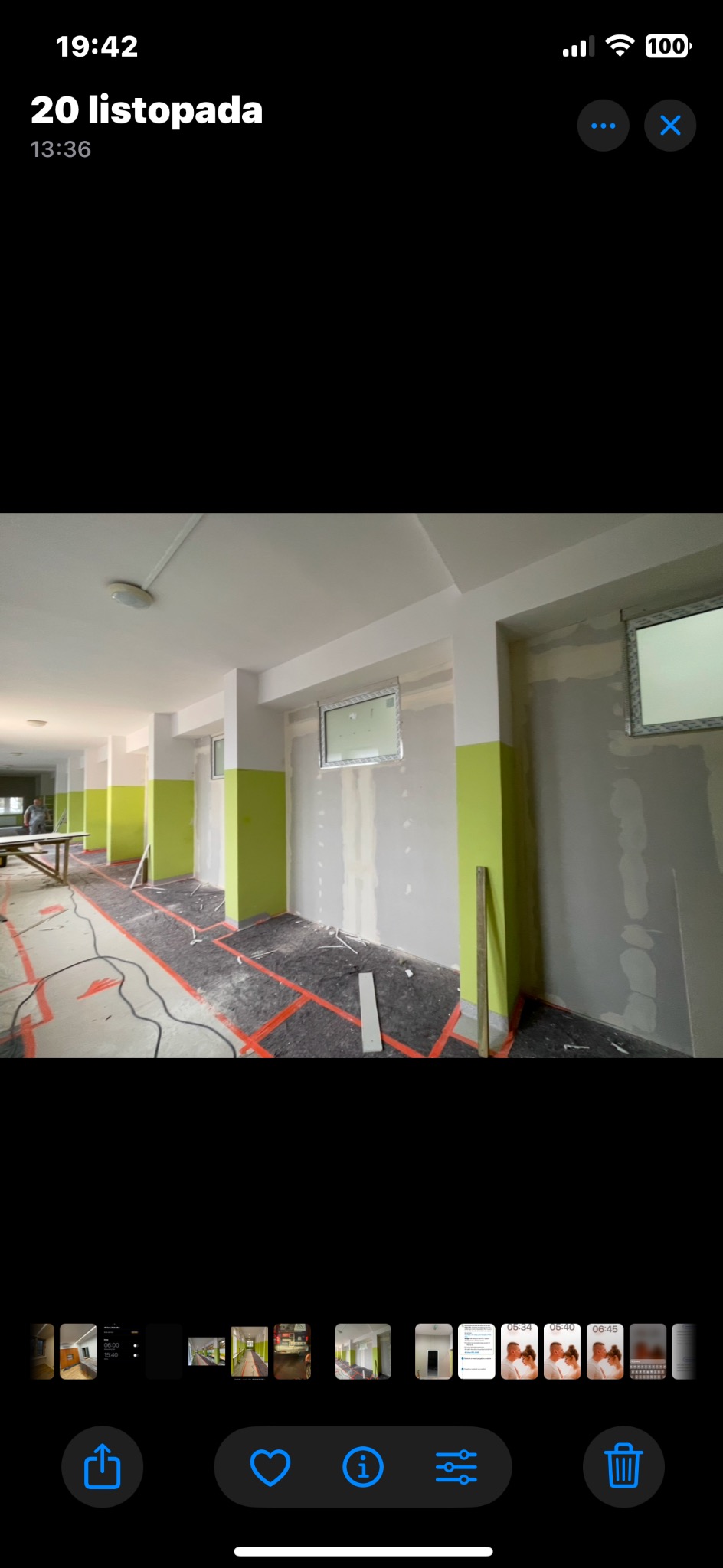 Korytarz w trakcie prac wykończeniowych: ściany częściowo pokryte szpachlą, na podłodze folia ochronna z pomarańczowymi liniami, widoczne pionowe pasy świeżo pomalowane na zielono. W tle widoczny...