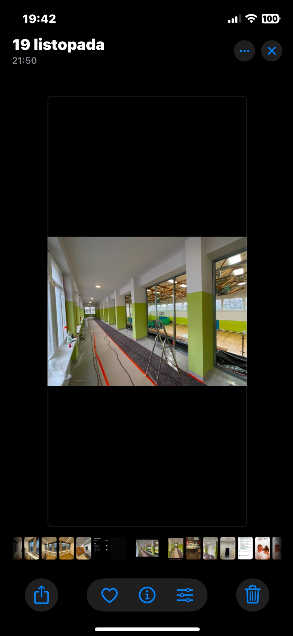 Długi korytarz w trakcie malowania, zabezpieczona podłoga, widoczne okna i drzwi do sali gimnastycznej.