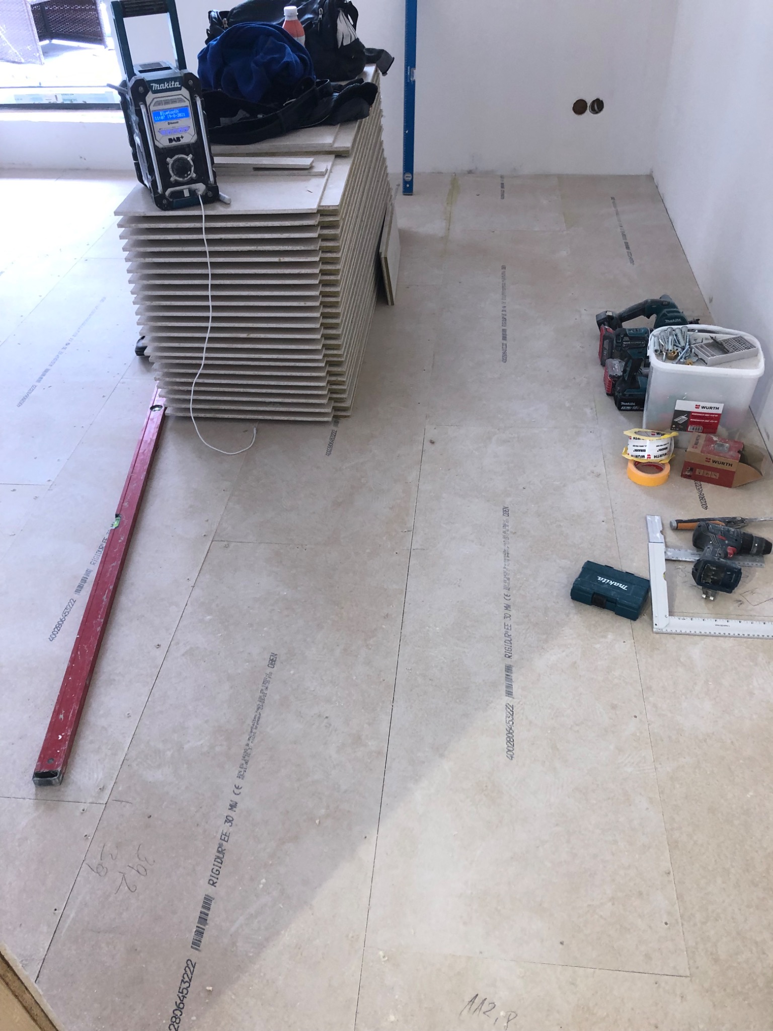 Stos płyt z widocznymi oznaczeniami producenta RIGIDUR, narzędzia budowlane, poziomica i radio Makita na podłodze w trakcie prac wykończeniowych.