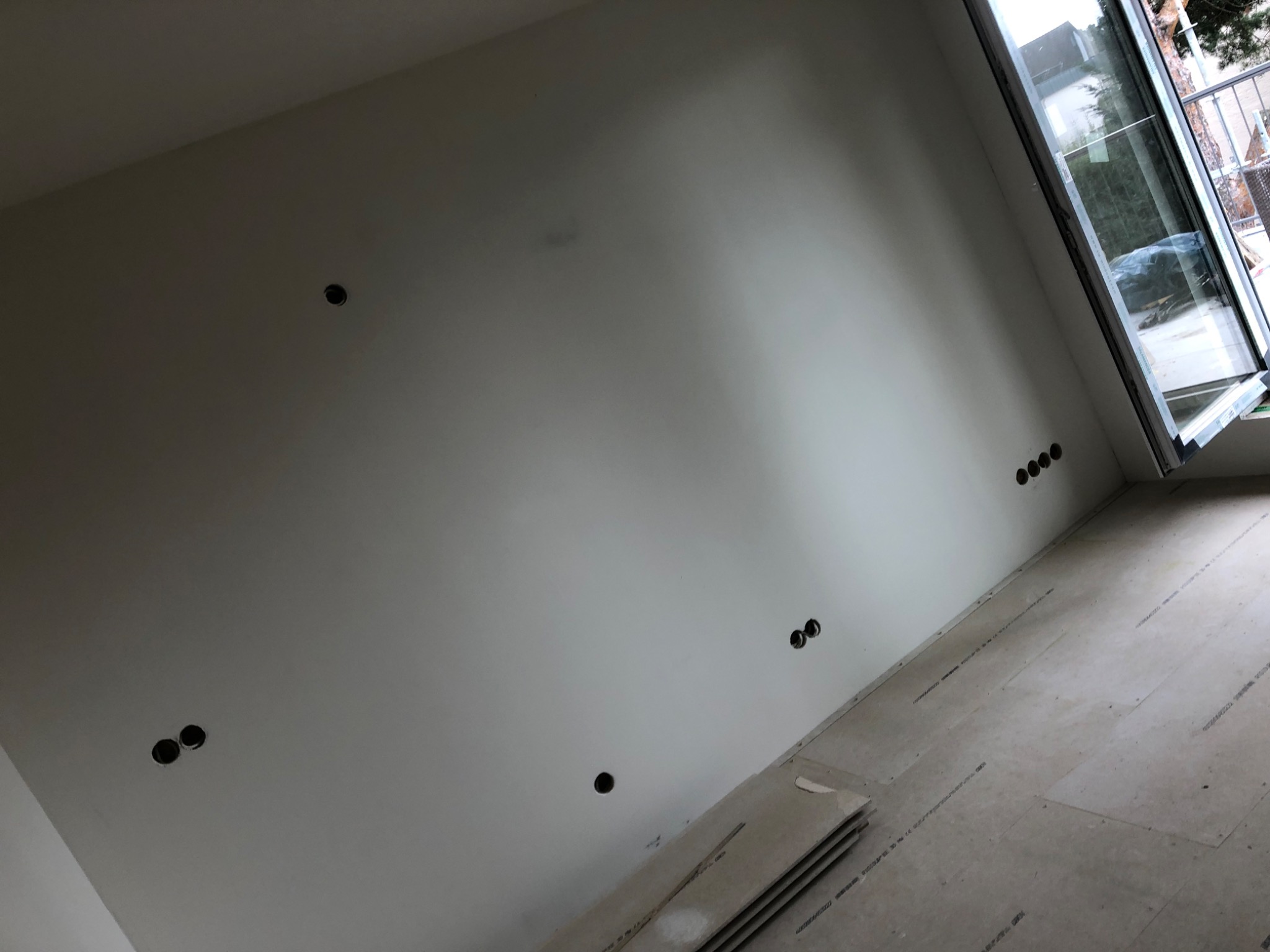 Ściana z wywierconymi otworami pod gniazdka elektryczne i oknem balkonowym, płyty gipsowe leżące na podłodze.