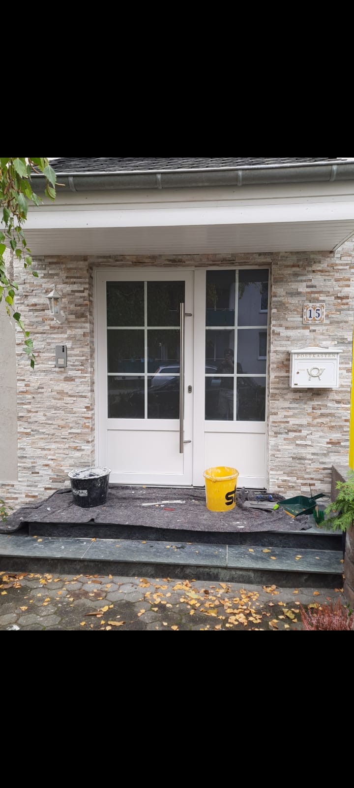 Białe, przeszklone drzwi wejściowe po montażu, z widocznymi dwoma wiaderkami po farbie i narzędziami na zabezpieczającej macie, osadzonymi w elewacji z kamienia dekoracyjnego.