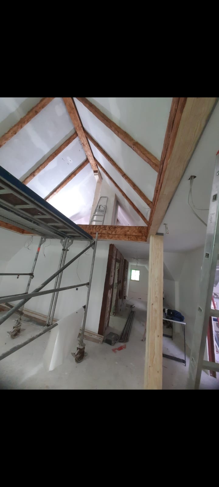 Poddasze w trakcie prac wykończeniowych: widoczne drewniane belki stropowe, częściowo zamontowane płyty gipsowo-kartonowe na skosach, rusztowanie i drabina.