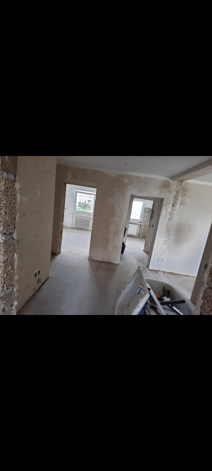 Wnętrze mieszkania w trakcie prac przygotowawczych do wykończenia, widoczne ściany po wstępnym wyrównaniu, podłoga wylewka, w tle okna i grzejnik, w pomieszczeniu prowizoryczny pojemnik na odpady...