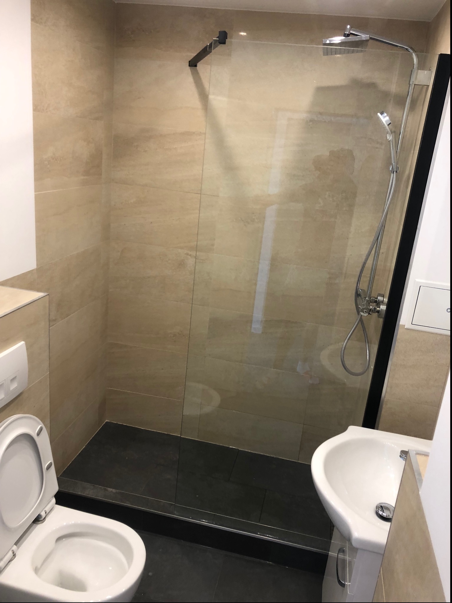 Wykończona łazienka z kabiną prysznicową typu walk-in z czarną ramą, umywalką na szafce i toaletą z otwartą deską, wykończona beżowymi płytkami imitującymi marmur i czarną podłogą.