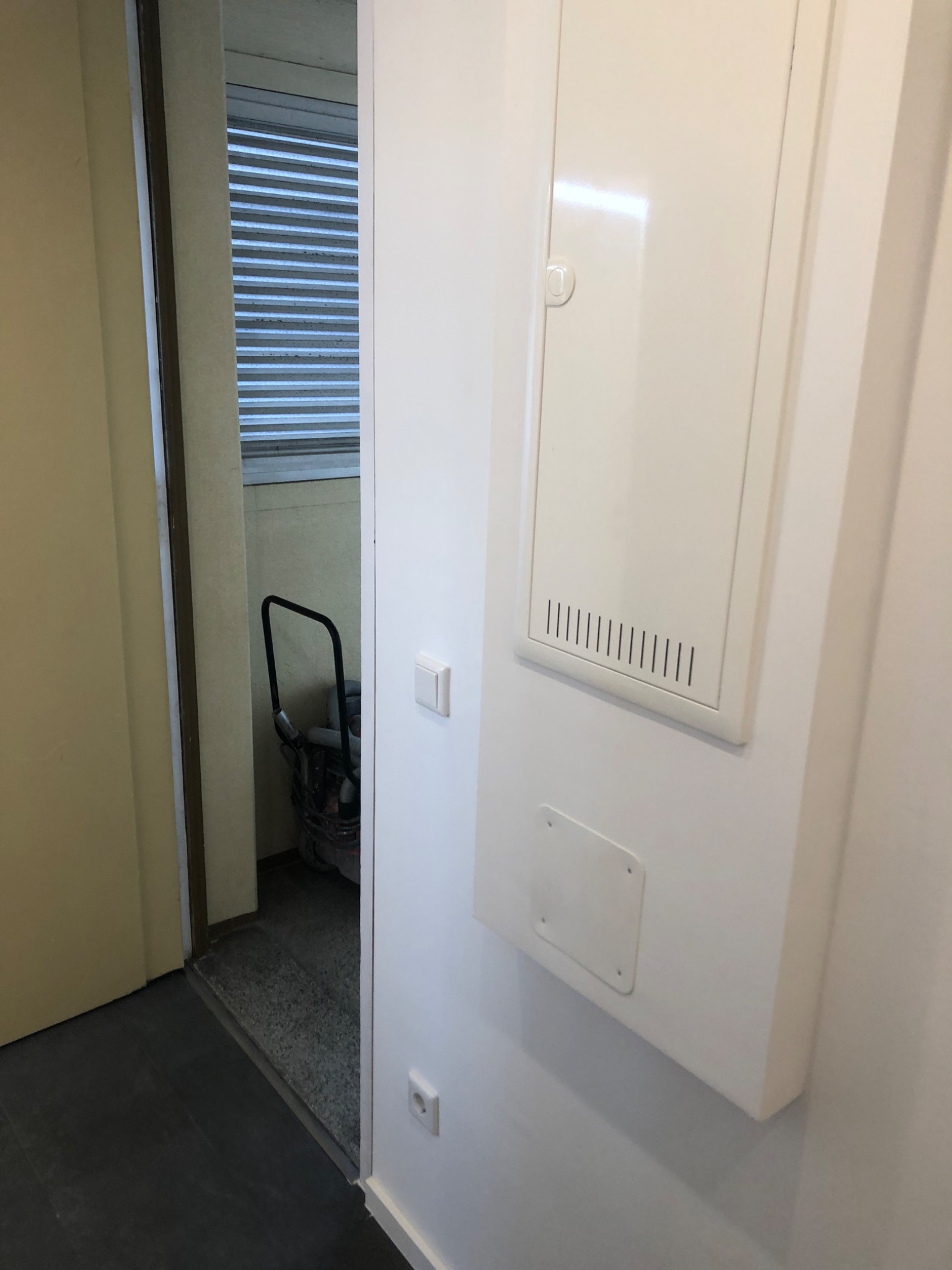 Biała skrzynka elektryczna na ścianie w korytarzu, widoczny przełącznik światła i gniazdko elektryczne, w tle widoczny sprzęt budowlany w pomieszczeniu z żaluzjami.
