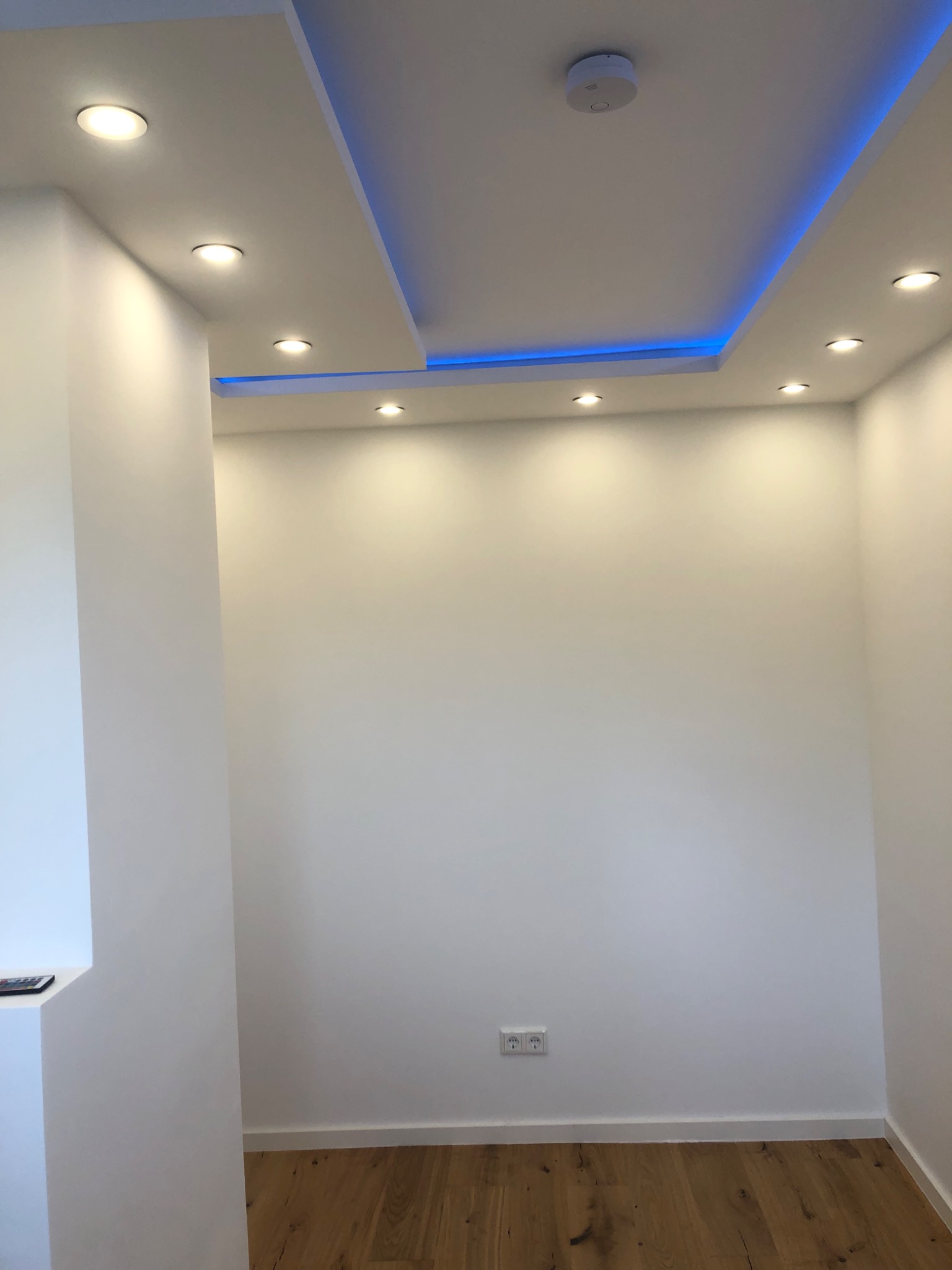 Nowoczesne wnętrze z białymi ścianami, drewnianą podłogą i podwieszanym sufitem z oświetleniem punktowym oraz niebieskim podświetleniem LED, widoczny czujnik dymu.