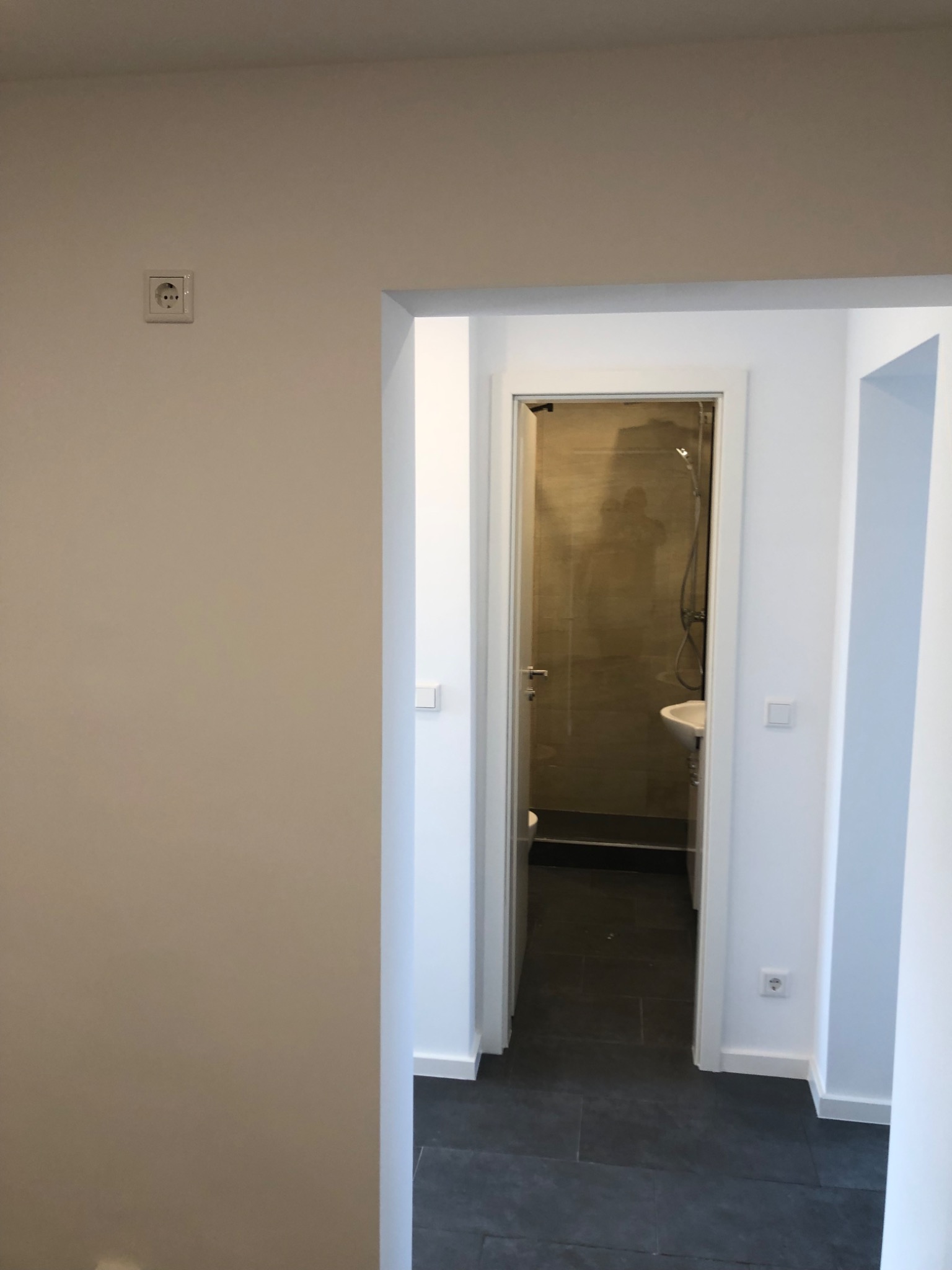 Wykończone wnętrze mieszkania z widokiem na łazienkę z kabiną prysznicową i umywalką, widoczne szare płytki podłogowe i białe ściany z gniazdkami elektrycznymi.
