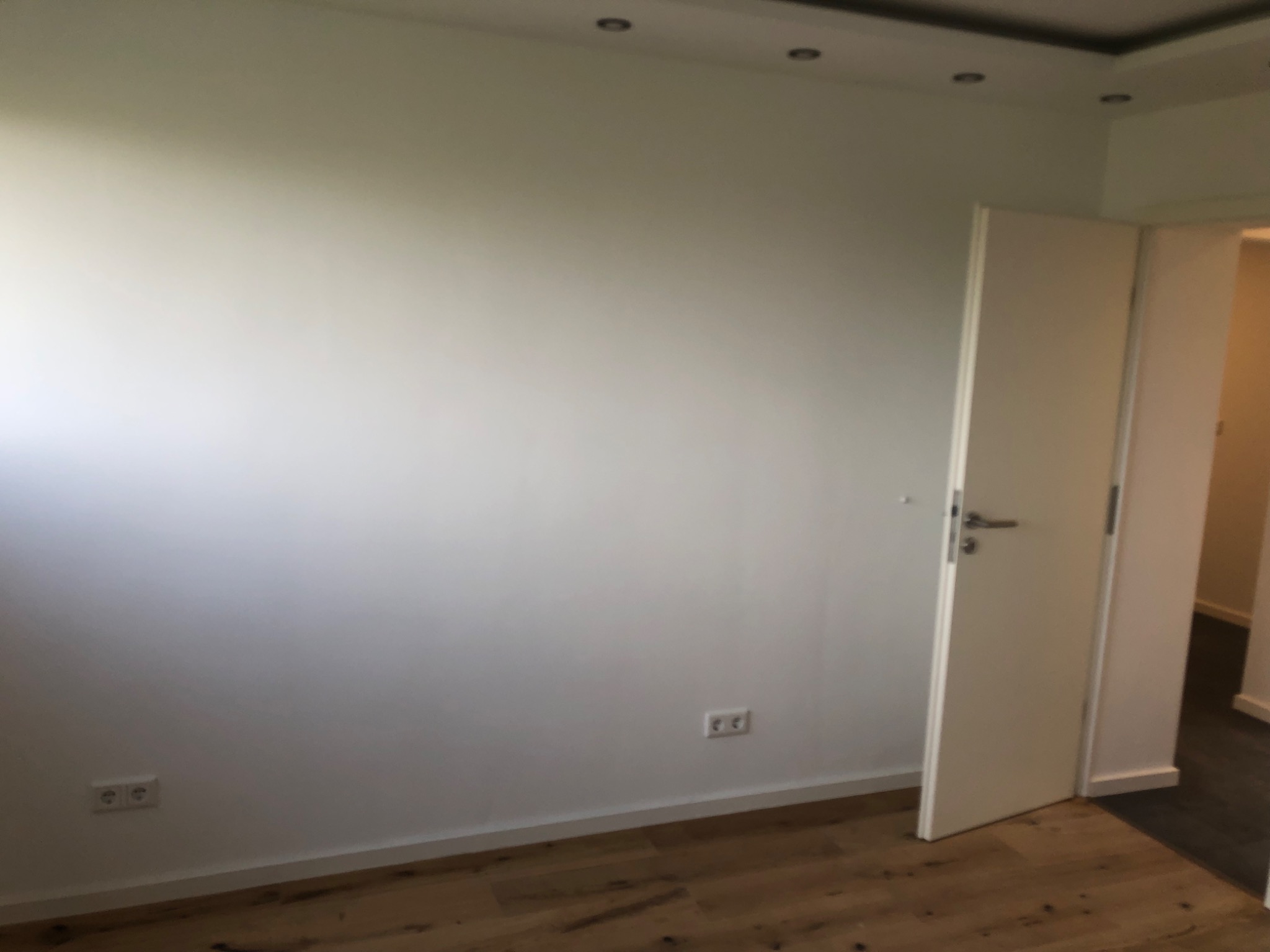 Wnętrze pokoju po remoncie z jasną ścianą, drewnianą podłogą, białymi drzwiami i widocznymi gniazdkami elektrycznymi.