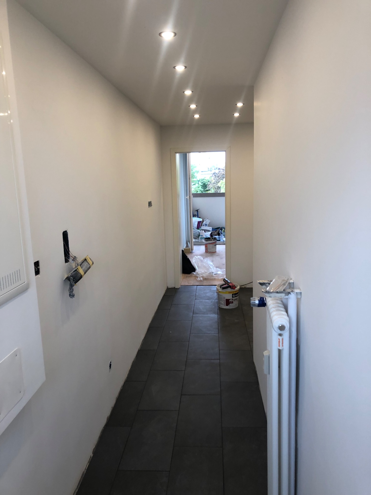 Korytarz w trakcie odświeżania, ściany pomalowane na biało, widoczne puszki elektryczne i grzejnik, na podłodze ciemne płytki, w tle otwarte drzwi na balkon.