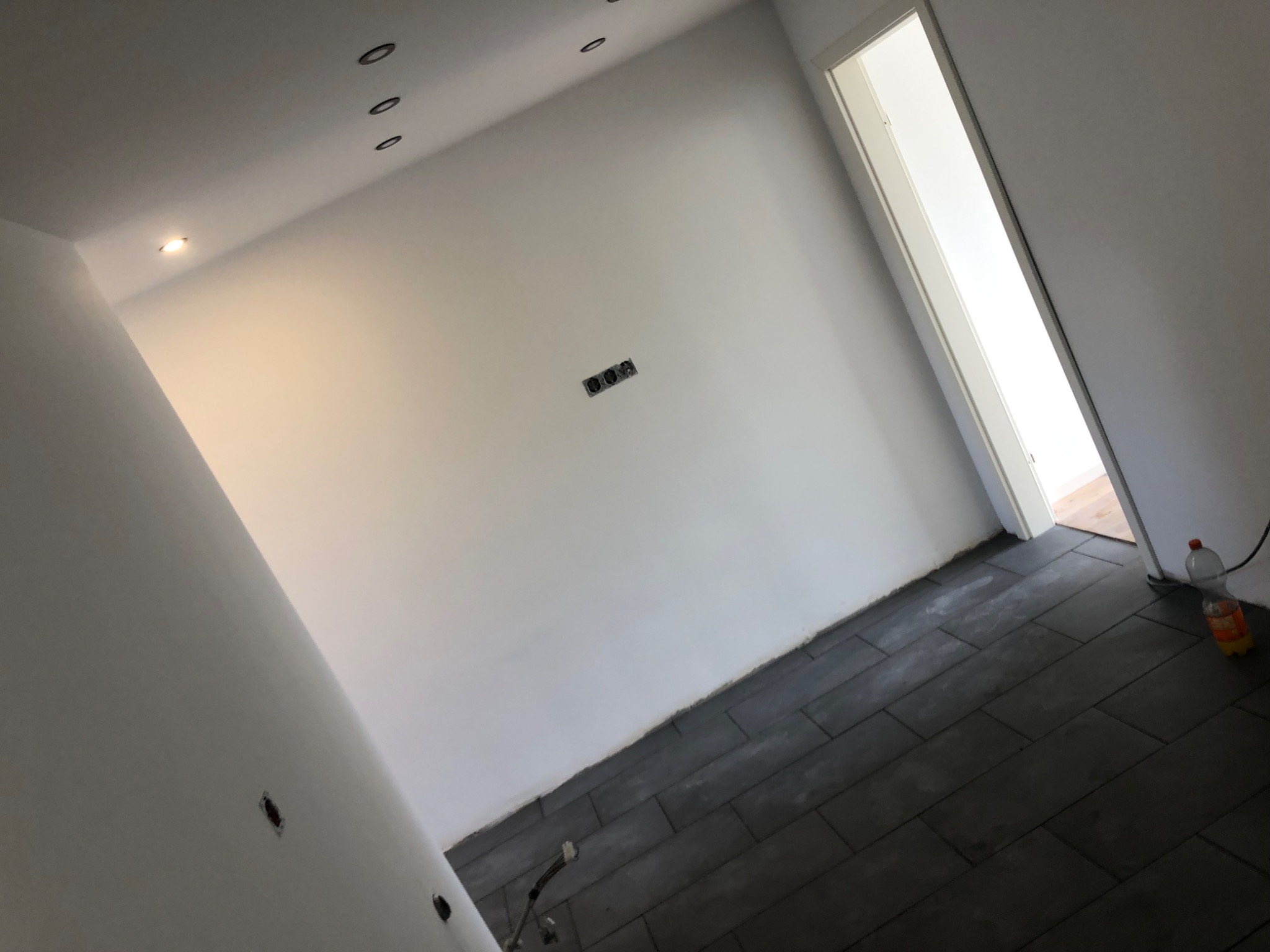 Wnętrze w trakcie wykańczania: ułożone ciemne panele podłogowe, białe ściany z widocznymi puszkami elektrycznymi, oświetlenie sufitowe i otwarte drzwi do sąsiedniego pomieszczenia.