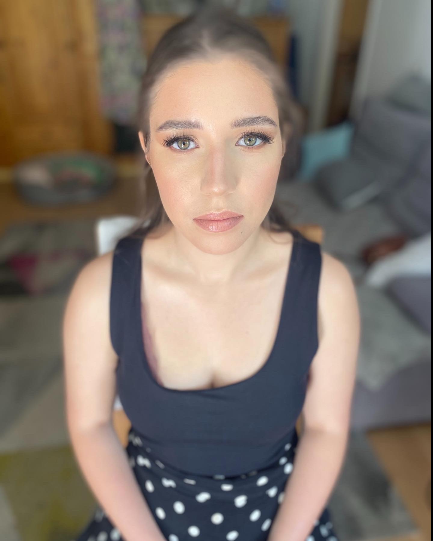 Portret kobiety z delikatnym makijażem, uwydatnione rzęsy, neutralne usta, spojrzenie skierowane wprost, na tle rozmytego wnętrza pokoju.