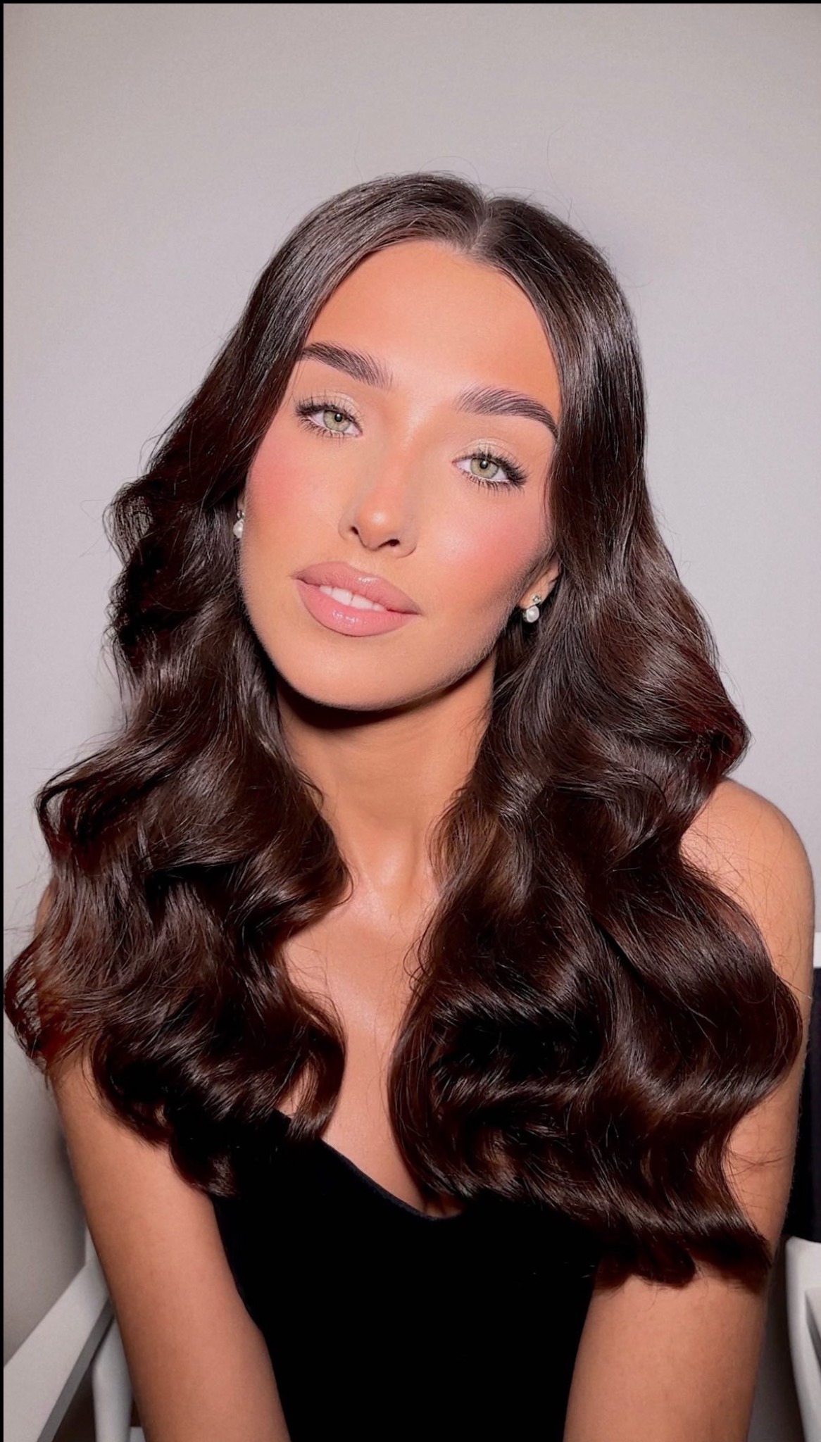 Portret kobiety z profesjonalnym makijażem, uwydatnione kości policzkowe różem, błyszczące powieki i usta, brązowe falowane włosy i perłowe kolczyki.