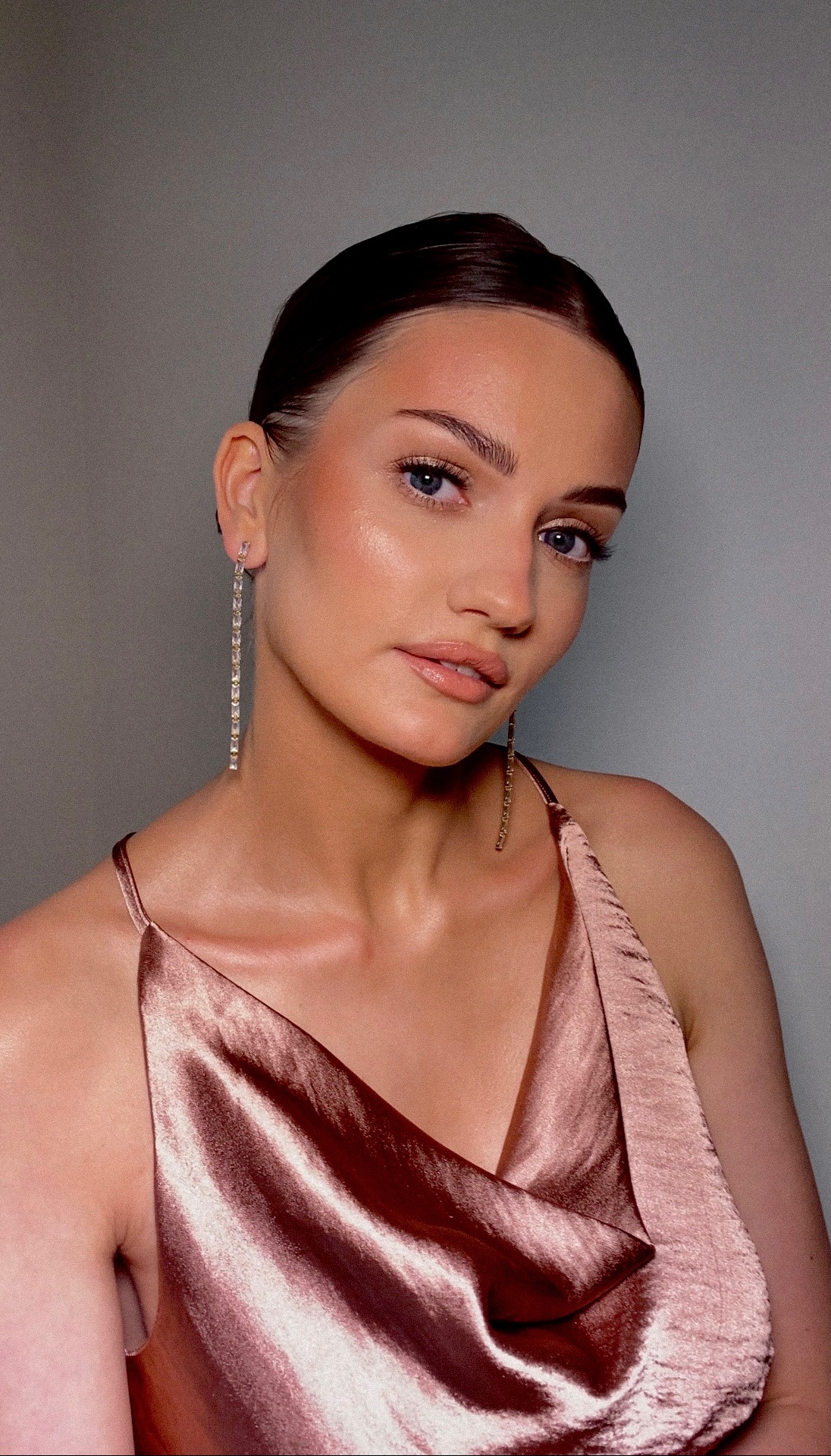 Portret kobiety z delikatnym, rozświetlającym wykończeniem, długie, błyszczące kolczyki i satynowa sukienka na ramiączkach.