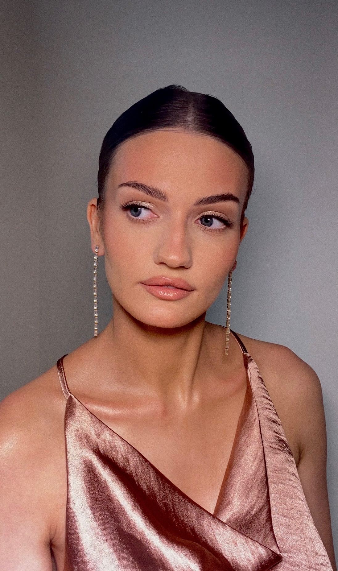 Portret kobiety z delikatnym, błyszczącym wykończeniem skóry, subtelnie podkreślonymi brwiami i długimi, kryształowymi kolczykami, ubrana w satynową sukienkę na ramiączkach.