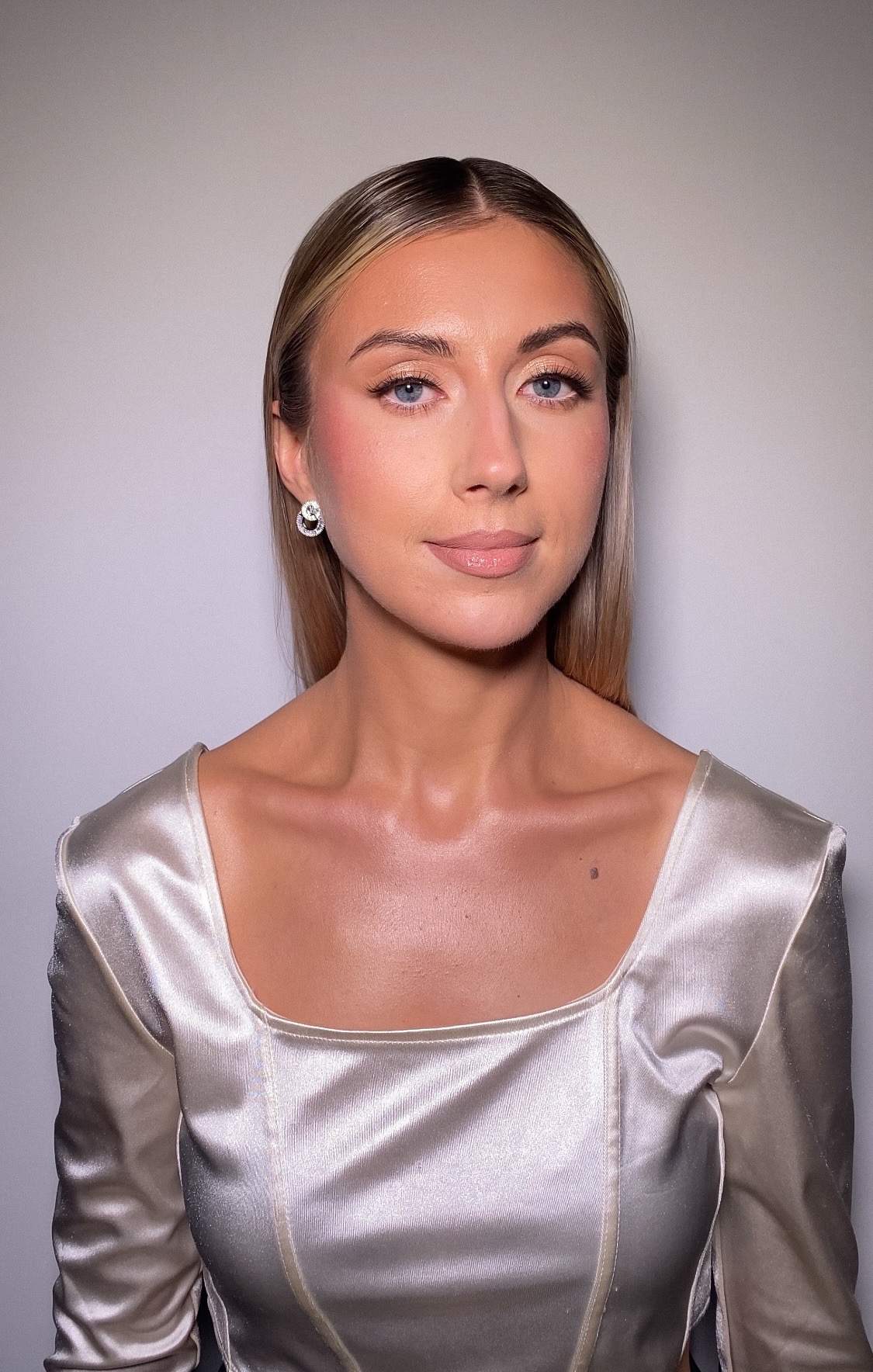 Portret kobiety o blond włosach i niebieskich oczach, z delikatnym rumieńcem i błyszczącymi ustami, ubrana w satynową bluzkę z kwadratowym dekoltem, eksponującą pieprzyk na szyi.