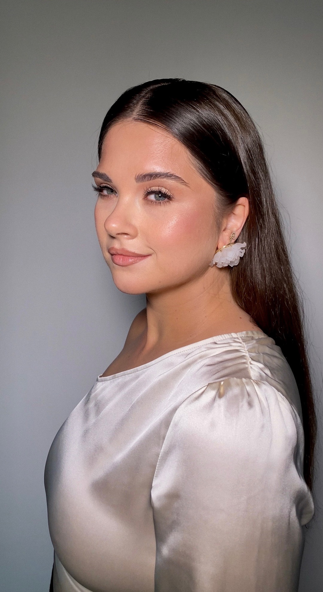 Portret kobiety z delikatnym, naturalnym upiększeniem, podkreślonymi rzęsami i błyszczącą cerą, ubrana w satynową bluzkę i ozdobne kolczyki.