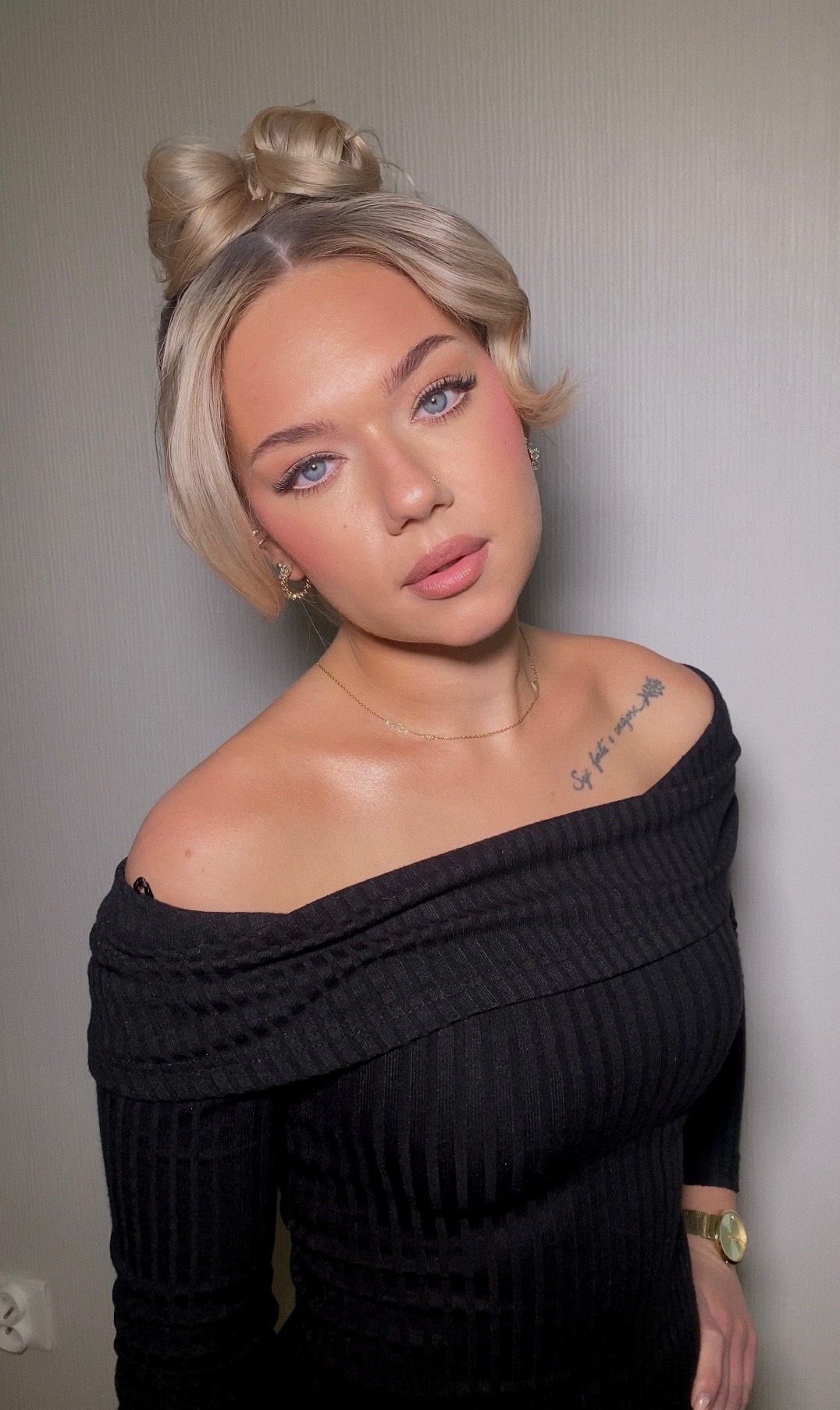Portret młodej kobiety z blond włosami upiętymi w kok, delikatny róż na policzkach i subtelnie podkreślone oczy, widoczny tatuaż na obojczyku.