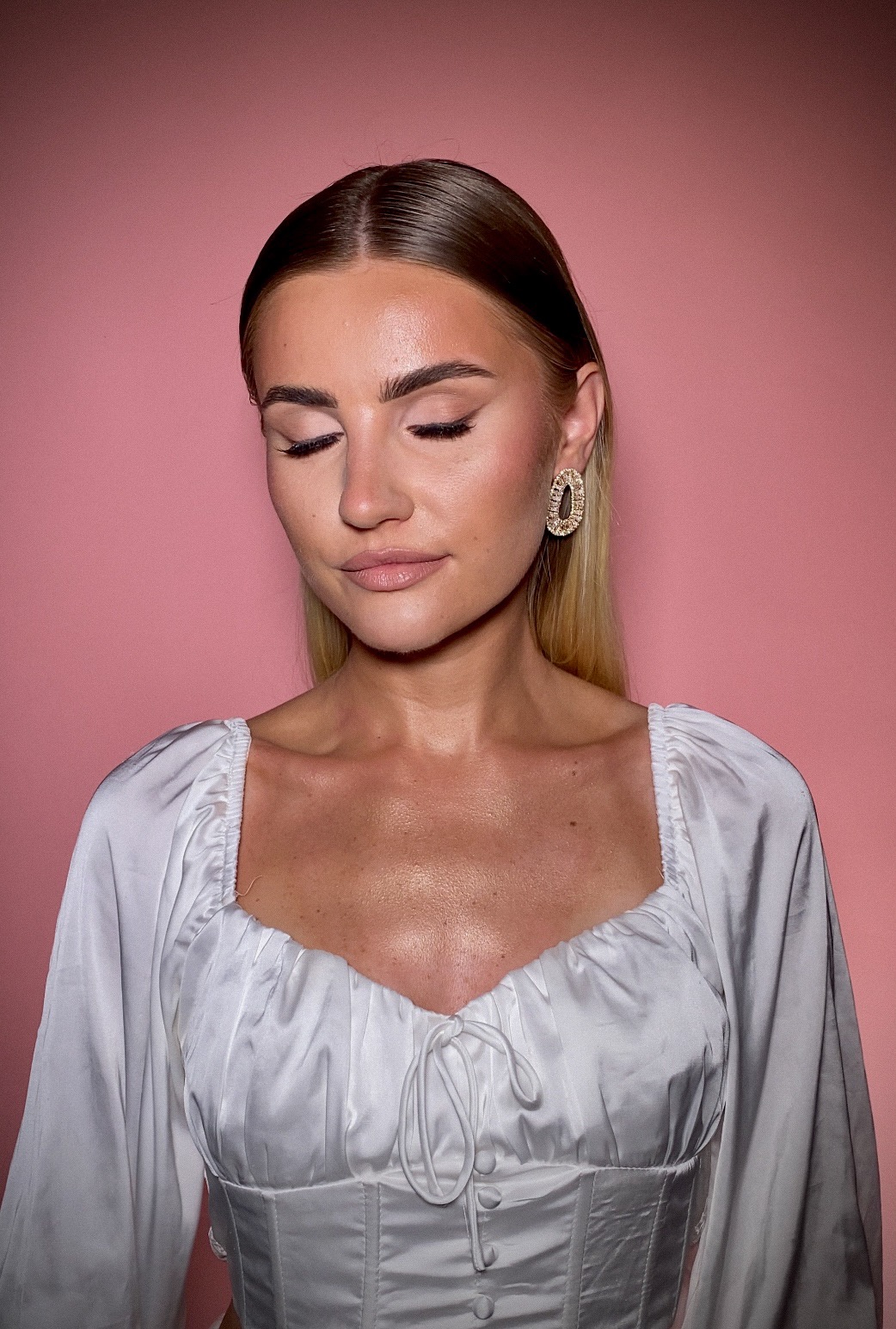 Portret kobiety z zamkniętymi oczami, delikatny kontur oka, błyszcząca skóra, złote kolczyki na tle różowego studia.