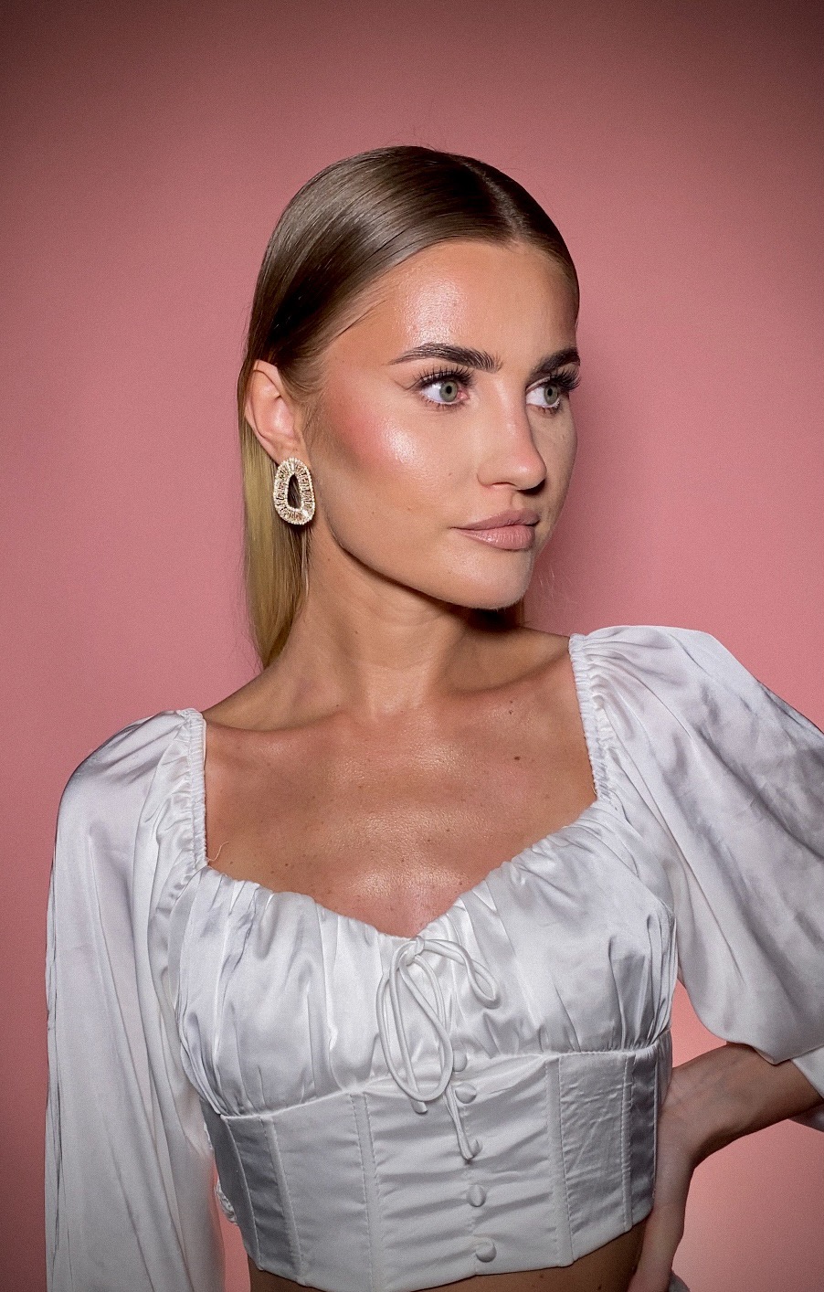 Portret kobiety z delikatnym makijażem, błyszczącą skórą, widocznym różem na policzkach i dużymi złotymi kolczykami na tle różowego studia.