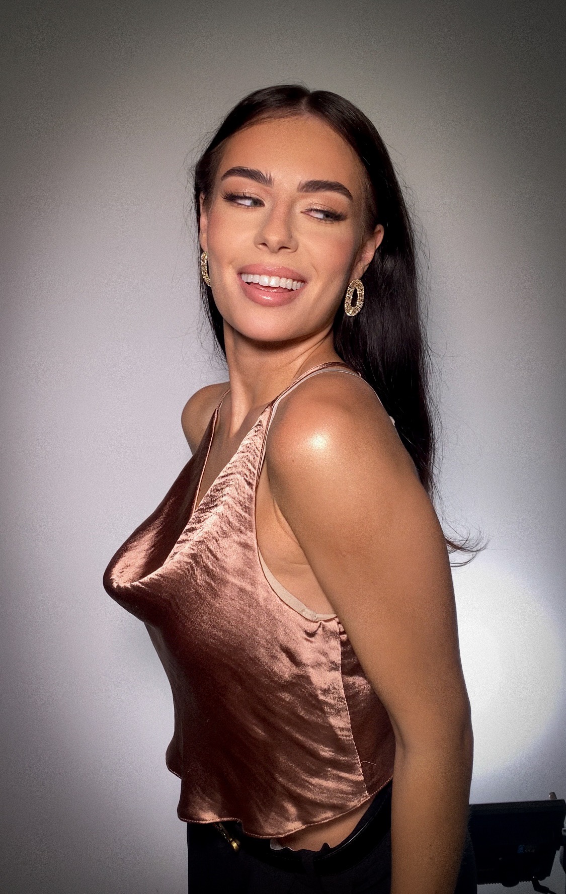 Uśmiechnięta brunetka z błyszczącym makijażem, w satynowym topie i złotych kolczykach, prezentuje profesjonalny makijaż.