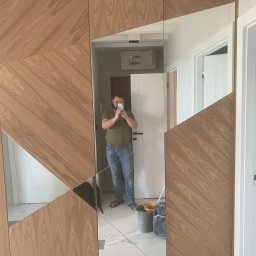 REMONTY - WYKOŃCZENIA ARTEM - PROJEKT RAFAŁ WIŚNIEWSKI - Nowoczesna szafa wnękowa z drewnianymi panelami ułożonymi w geometryczny wzór, z lustrzanymi wstawkami, odbijająca wnętrze mieszkania i mężczyznę robiącego zdjęcie telefonem.