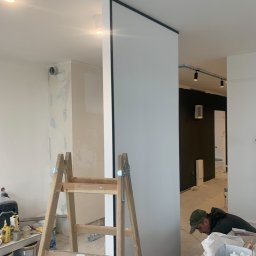 REMONTY - WYKOŃCZENIA ARTEM - PROJEKT RAFAŁ WIŚNIEWSKI - Wnętrze w trakcie prac wykończeniowych: widoczna drewniana drabina, częściowo wykończona ściana z czarną obwódką, pracownik w tle. Ściana przygotowana do malowania, widoczne ślady szpachlowania.