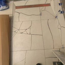 REMONTY - WYKOŃCZENIA ARTEM - PROJEKT RAFAŁ WIŚNIEWSKI - Układanie płytek podłogowych imitujących spękania w łazience, widoczne narzędzia glazurnicze, poziomica i krzyżyki dystansowe.