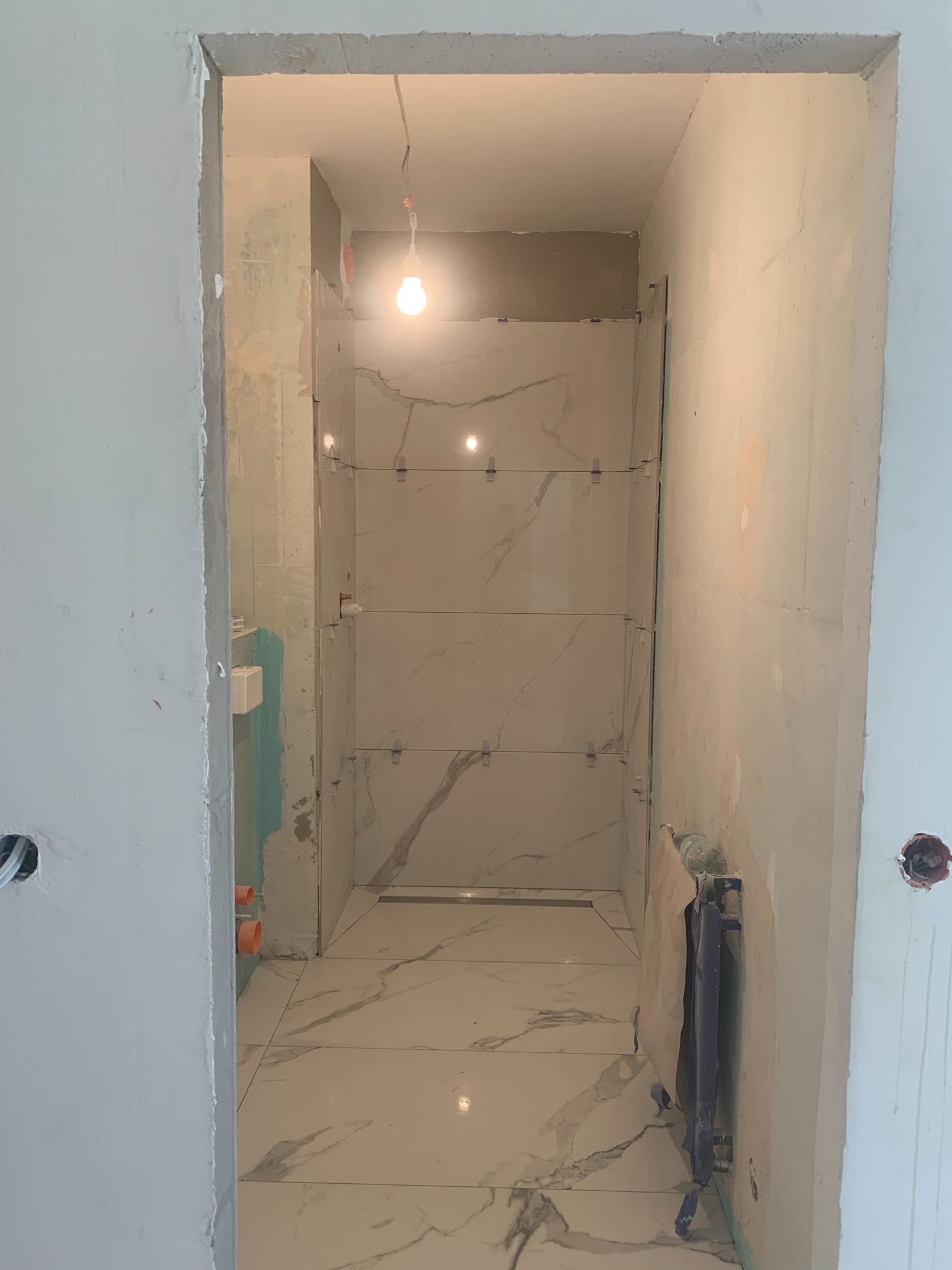 Wnętrze łazienki w trakcie prac wykończeniowych: ułożone płytki imitujące marmur na podłodze i ścianach, widoczne listwy dystansowe, surowa ściana z instalacją elektryczną, wisząca żarówka...