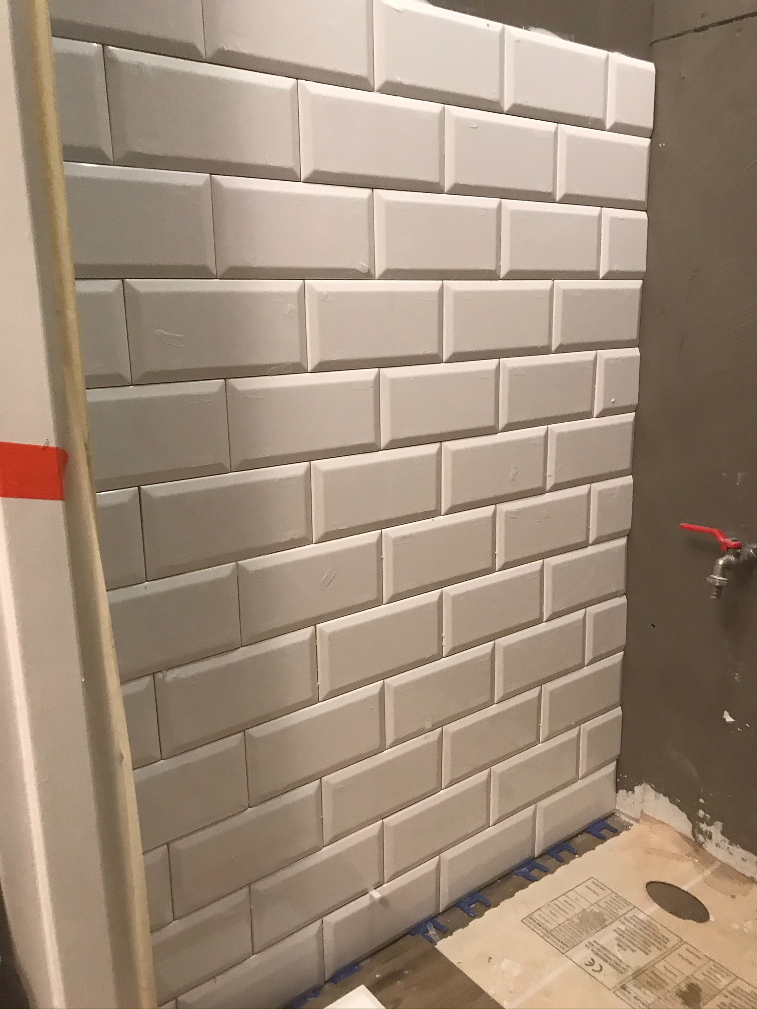 Ściana wyłożona białymi, wypukłymi płytkami typu metro, widoczne krzyżyki dystansowe u dołu, surowa ściana obok z kranem z czerwoną rączką.