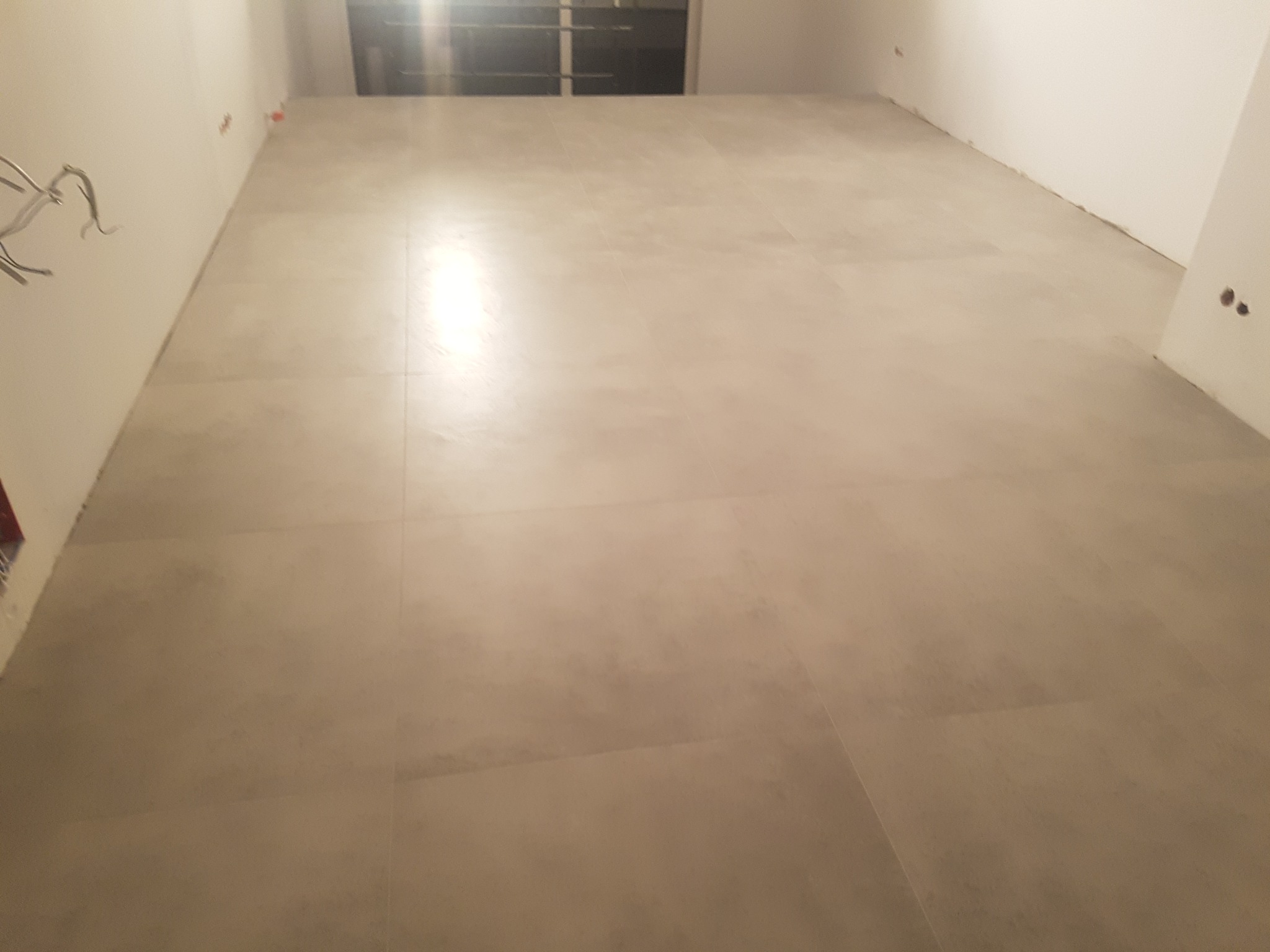 Jasnoszare płytki podłogowe ułożone w nowym pomieszczeniu, widoczne wystające kable ze ściany i niedokończone wykończenie ścian.