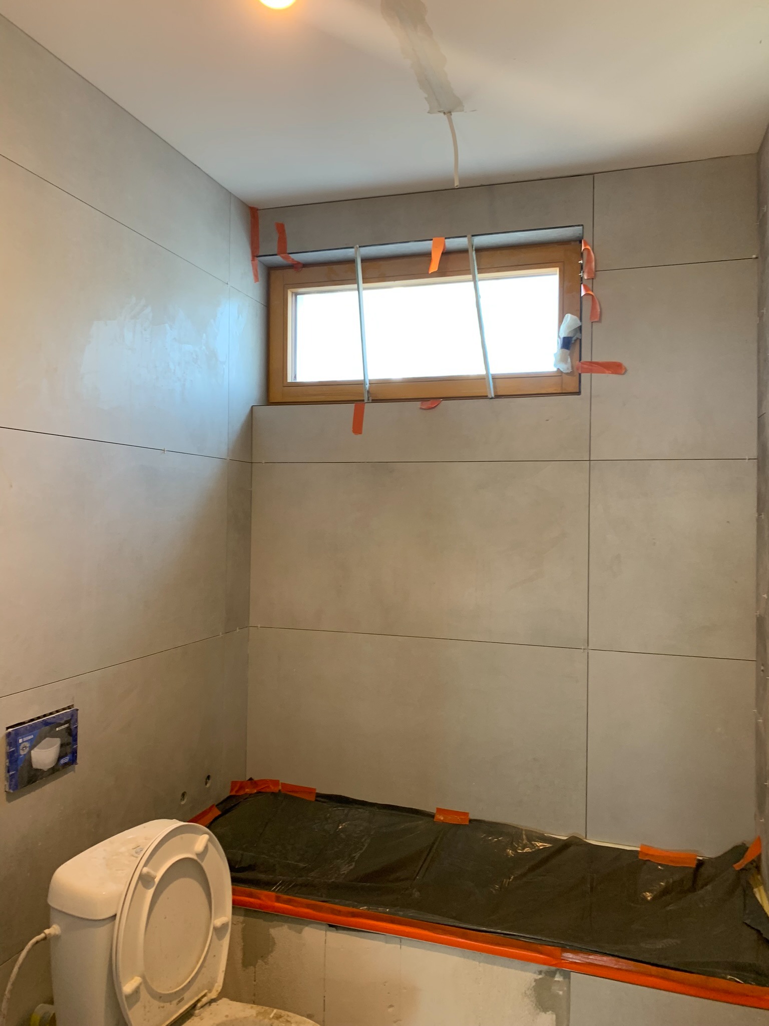 Wnętrze łazienki w trakcie kładzenia płytek ceramicznych w odcieniach szarości, zabezpieczona folią wanna, widoczne narzędzia glazurnicze i częściowo zamontowana toaleta.