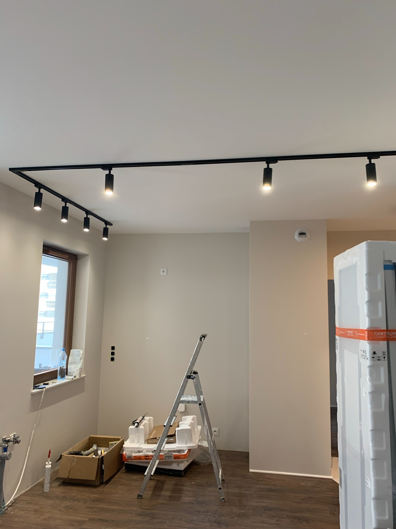 Montaż szynoprzewodu z reflektorami w nowym mieszkaniu w Warszawie, widoczna drabina, kartony z materiałami budowlanymi i instalacja wodna.