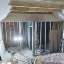 Strop z drewnianymi belkami, metalowy stelaż pod płyty karton-gips z izolacją z wełny mineralnej, widoczne narzędzia i reflektor na statywie w trakcie remontu poddasza.