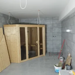 Montaż sauny w łazience z szarymi płytkami, widoczne narzędzia i materiały budowlane, niedokończone podłączenia elektryczne.