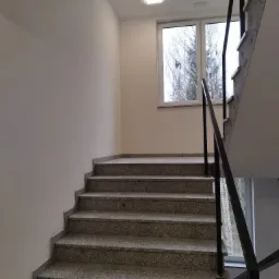 Klatka schodowa z szarymi granitowymi stopniami i czarną metalową balustradą, widok na okno z drzewami bez liści w tle.