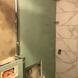 Montaż stelaża podtynkowego w łazience, widoczna płyta karton gips, metalowa konstrukcja, rury instalacyjne i fragment ułożonych płytek imitujących marmur.