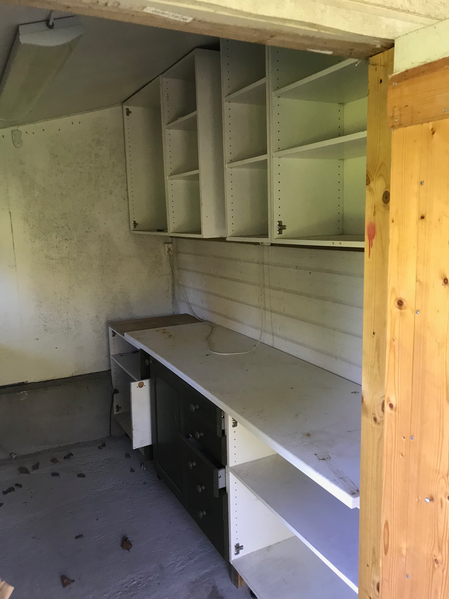 Wnętrze pomieszczenia gospodarczego przed remontem: widoczne stare, białe szafki wiszące i stojące, brudna podłoga i ściany z widocznymi plamami wilgoci, surowy drewniany element konstrukcyjny po...