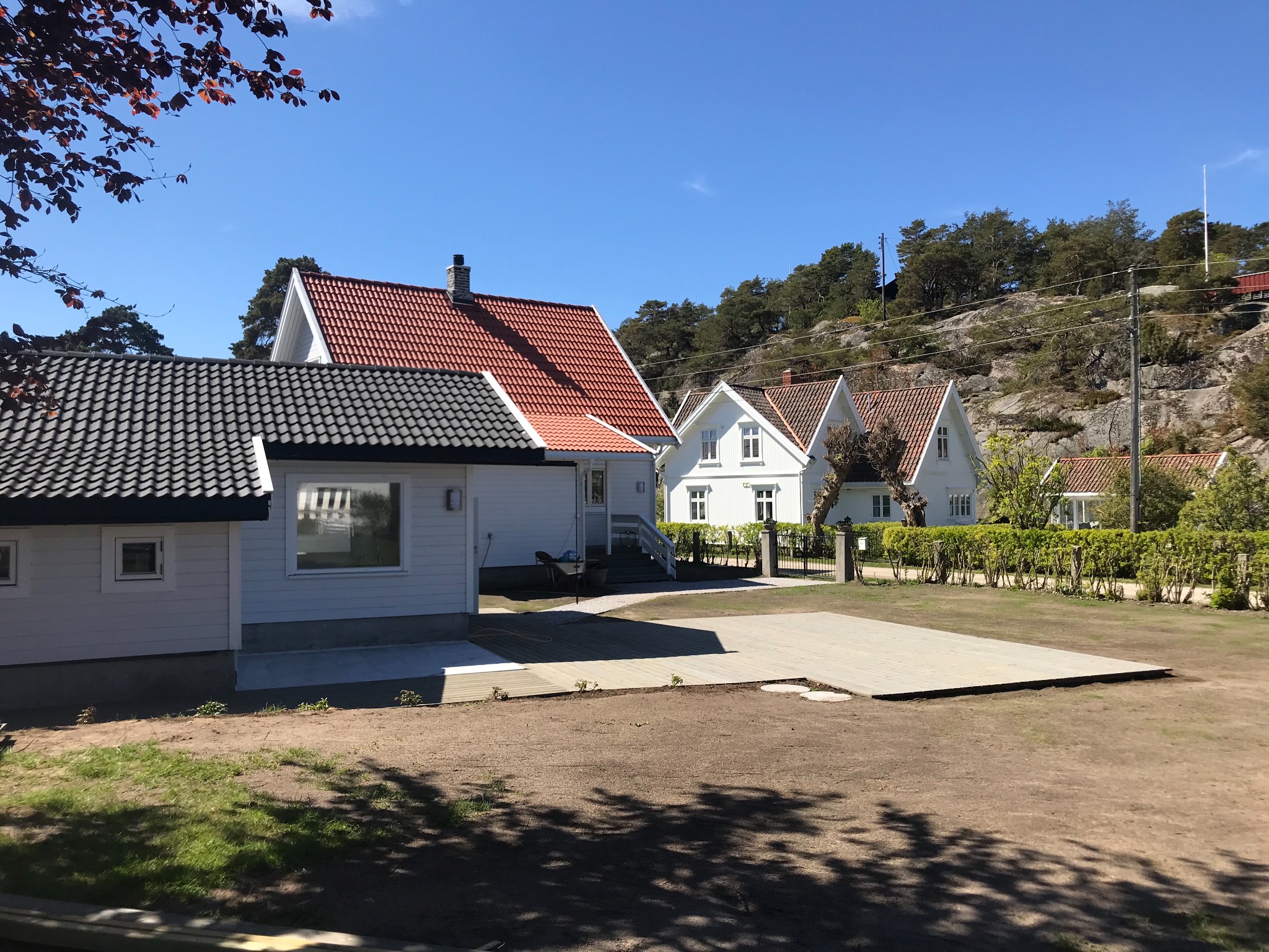 Nowo wybudowany drewniany taras przylegający do białego domu z ciemnoszarą dachówką, w tle inne domy z czerwonymi dachami i zalesione wzgórze, słoneczny dzień.