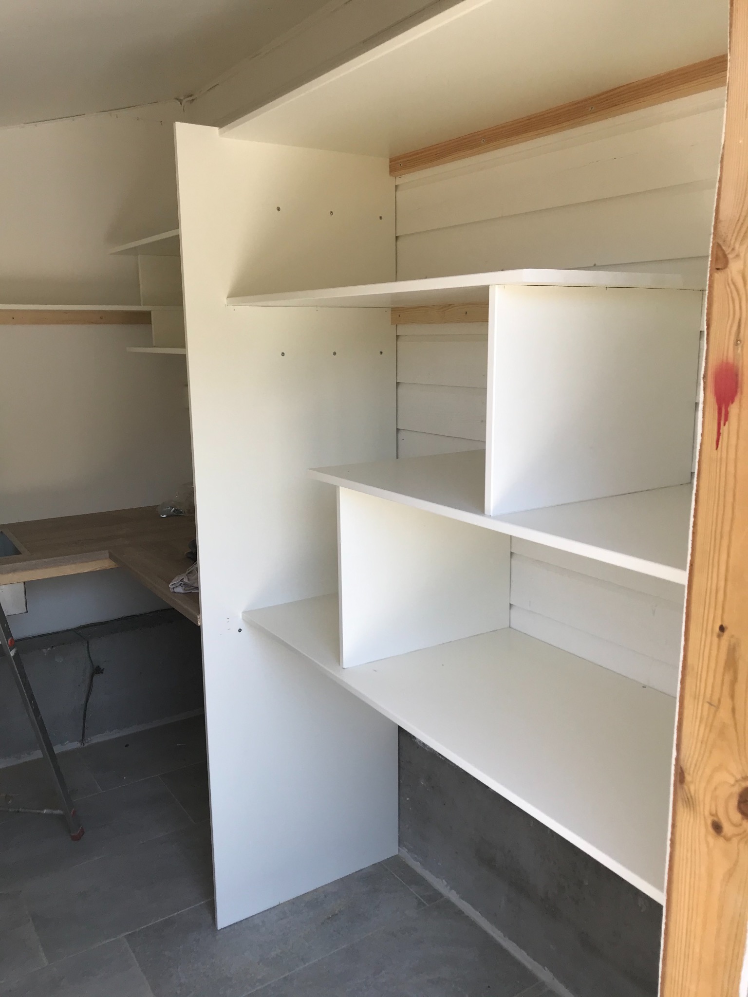 Wnętrze pomieszczenia w trakcie wykańczania z widoczną zabudową meblową z białych płyt, drewnianymi elementami konstrukcyjnymi i fragmentem drabiny opartym o ścianę.