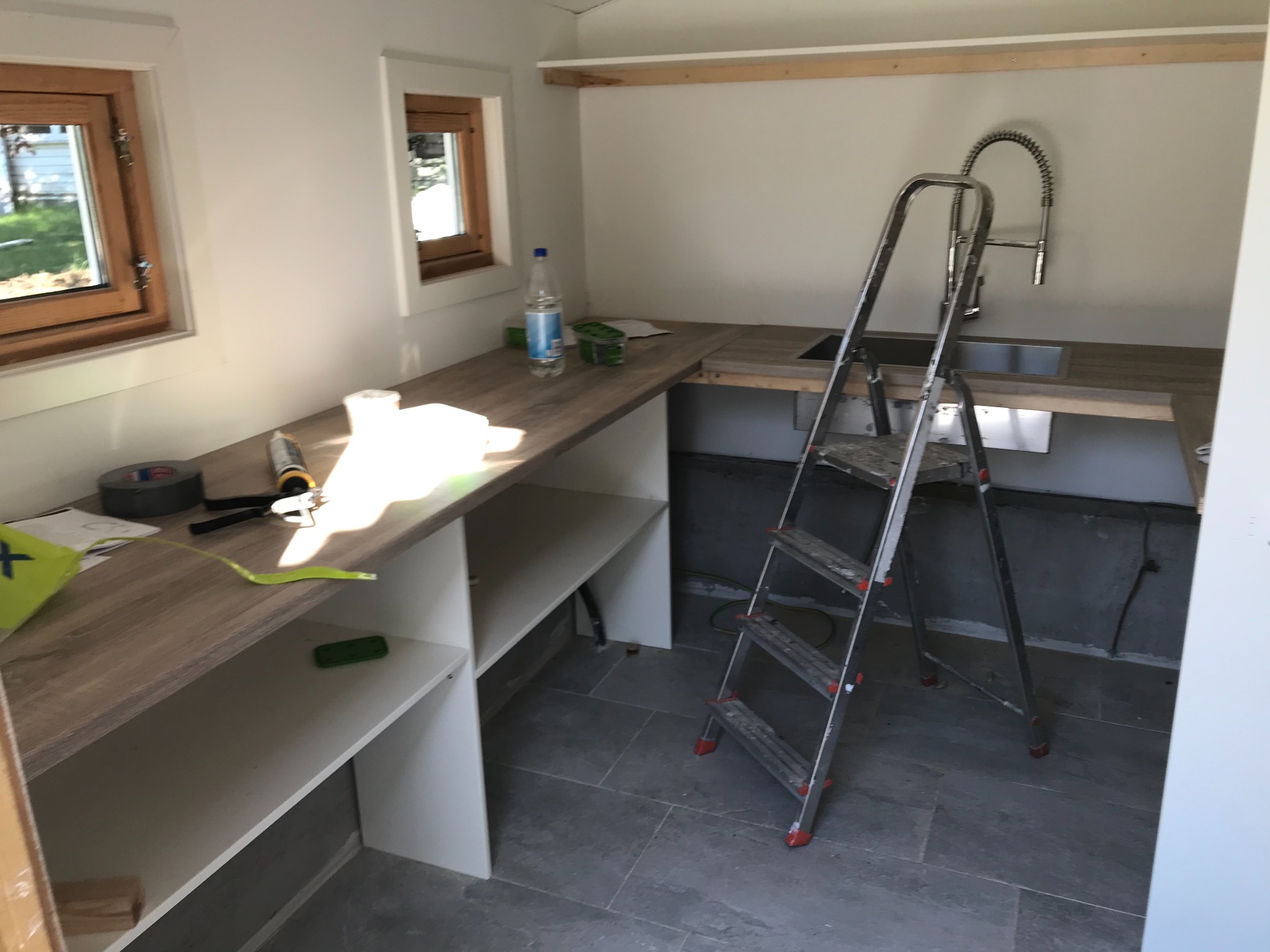 Wnętrze pomieszczenia w trakcie prac wykończeniowych: zamontowany blat z szafkami, widoczna drabina, zlew z baterią oraz narzędzia i materiały budowlane.
