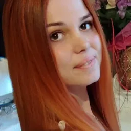 Portret młodej kobiety z długimi, rudymi włosami, delikatny uśmiech, w tle bukiet kwiatów i dekoracyjny przedmiot.