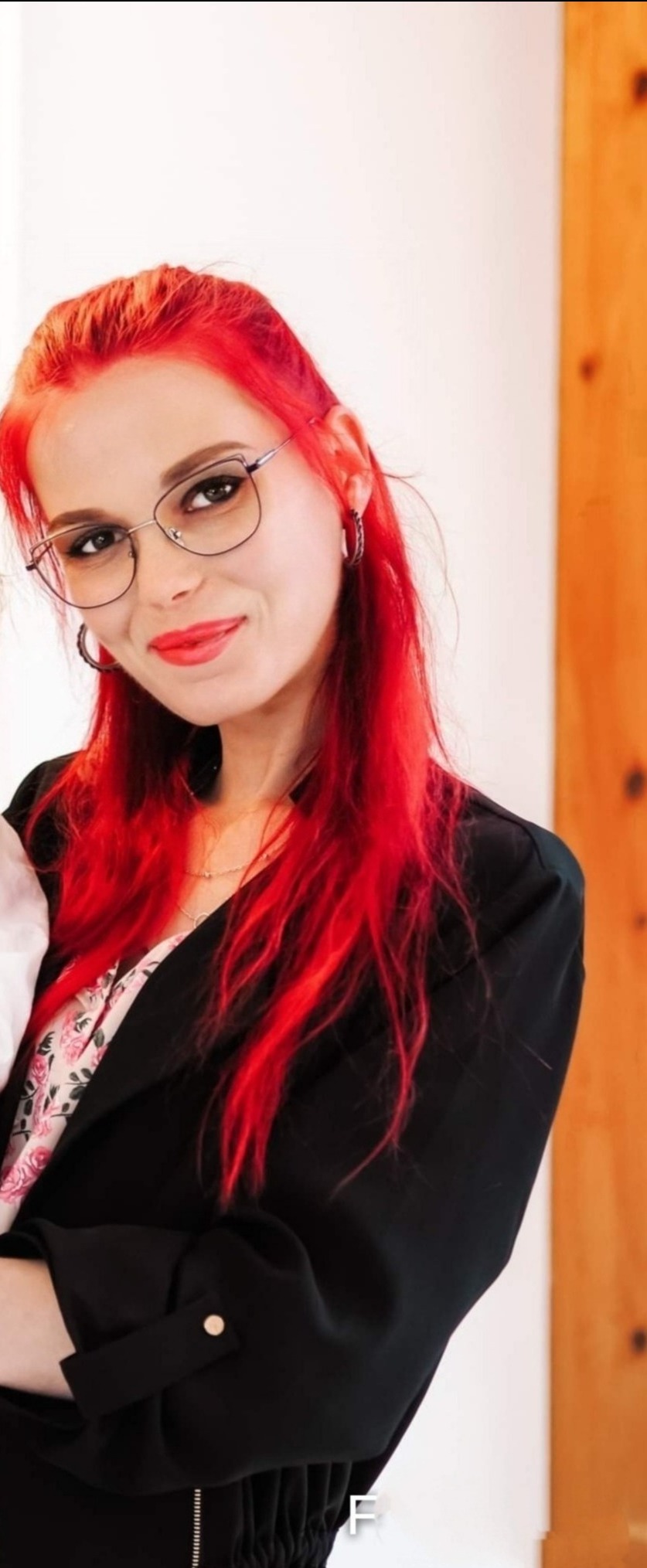 Portret uśmiechniętej kobiety z intensywnie czerwonymi włosami, w okularach o cienkich oprawkach i czarnej marynarce, z delikatnym makijażem ust.