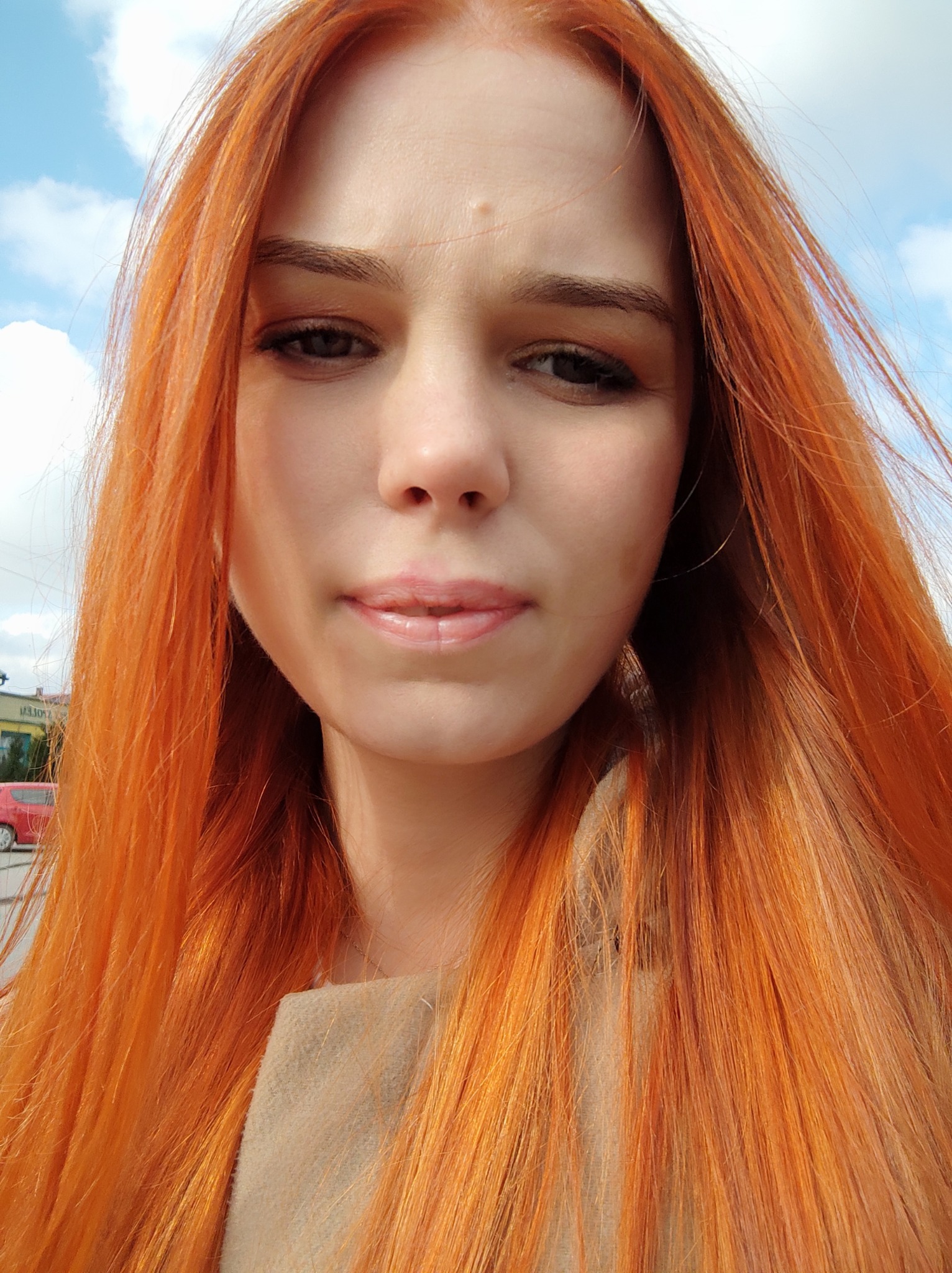 Portret kobiety z długimi, rudymi włosami, delikatny makijaż, widok z góry, w tle błękitne niebo z chmurami.