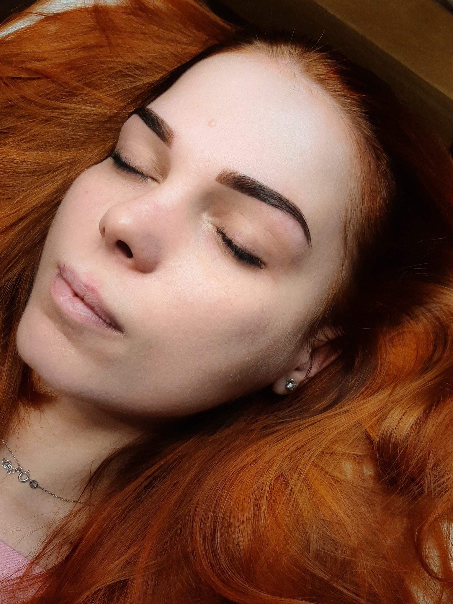 Portret kobiety z rudymi włosami, z zamkniętymi oczami i świeżo wyregulowanymi, ciemnymi brwiami, leżącej na ciemnym tle.