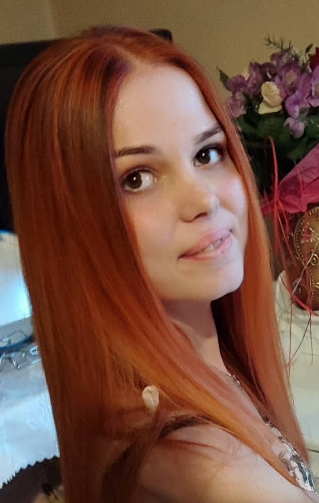 Portret młodej kobiety z długimi, rudymi włosami, delikatny uśmiech, w tle bukiet kwiatów i dekoracyjny przedmiot.