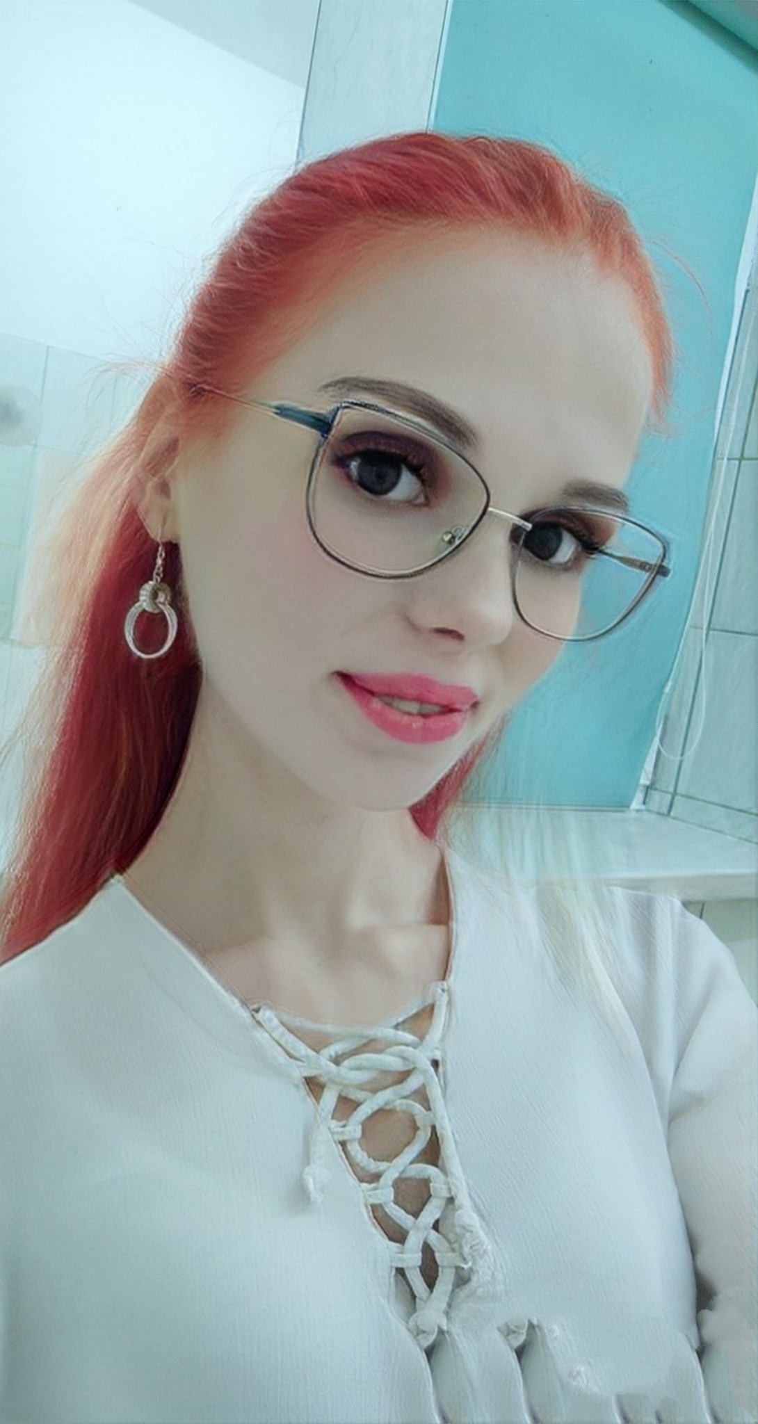 Portret kobiety z rudymi włosami i okularami w srebrnej oprawce, ubrana w białą bluzkę z wiązaniem na dekolcie, pozująca na tle błękitnej ściany.