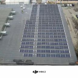 Widok z drona na rozległą instalację paneli słonecznych na płaskim dachu budynku przemysłowego, z widocznymi elementami wentylacji i fragmentem parkingu w tle.