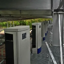 Szereg szarych jednostek zewnętrznych pomp ciepła na metalowej platformie z widokiem na zalesiony teren w tle.