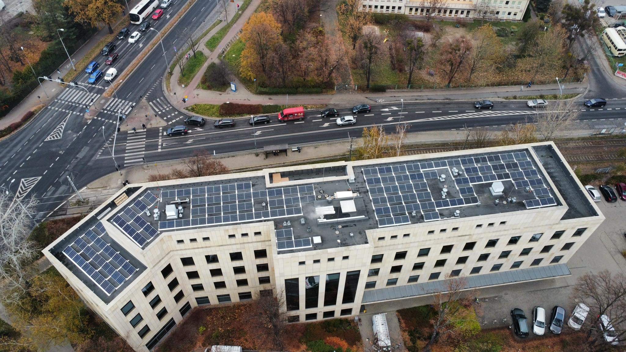 Widok z góry na budynek biurowy z panelami słonecznymi na dachu, ruchliwa ulica z przejściem dla pieszych i jesienne drzewa w tle.