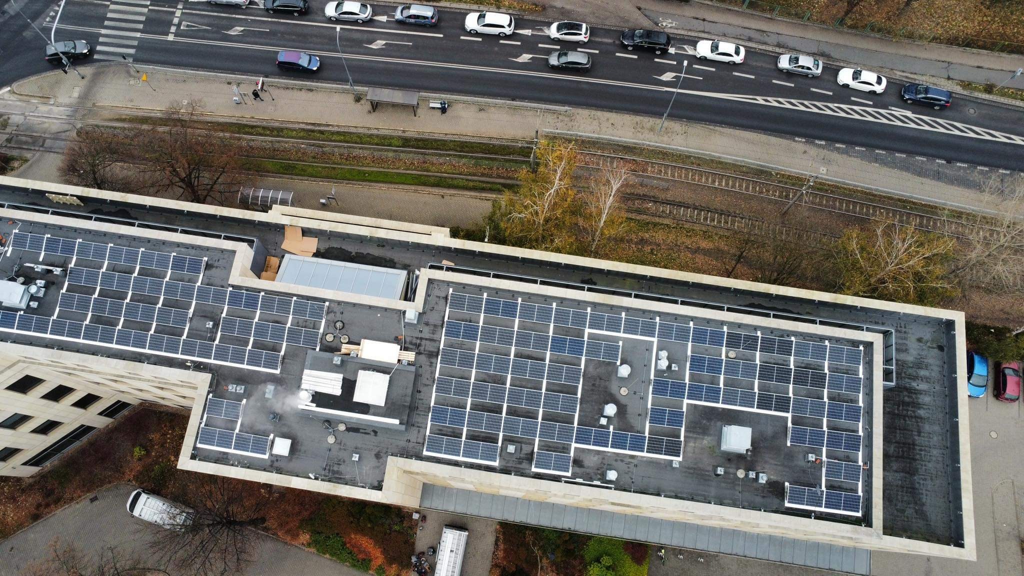 Widok z góry na budynek z zainstalowanymi panelami słonecznymi na dachu, obok biegnie ulica z samochodami i linia kolejowa.